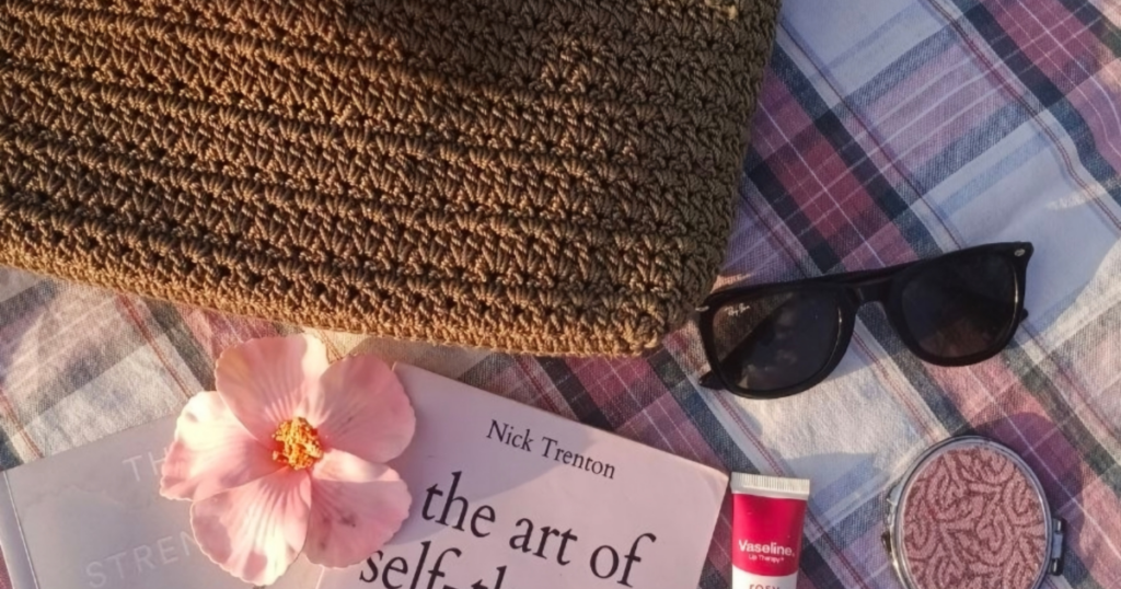 sunglasses, a woven tote and a book with a pink flower in the style of a flat lay